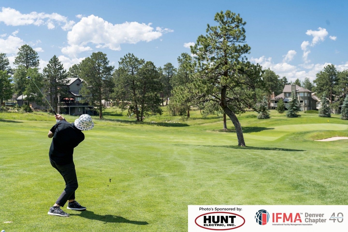 Golfer swinging club on a green course, trees and houses in the background under a blue sky.