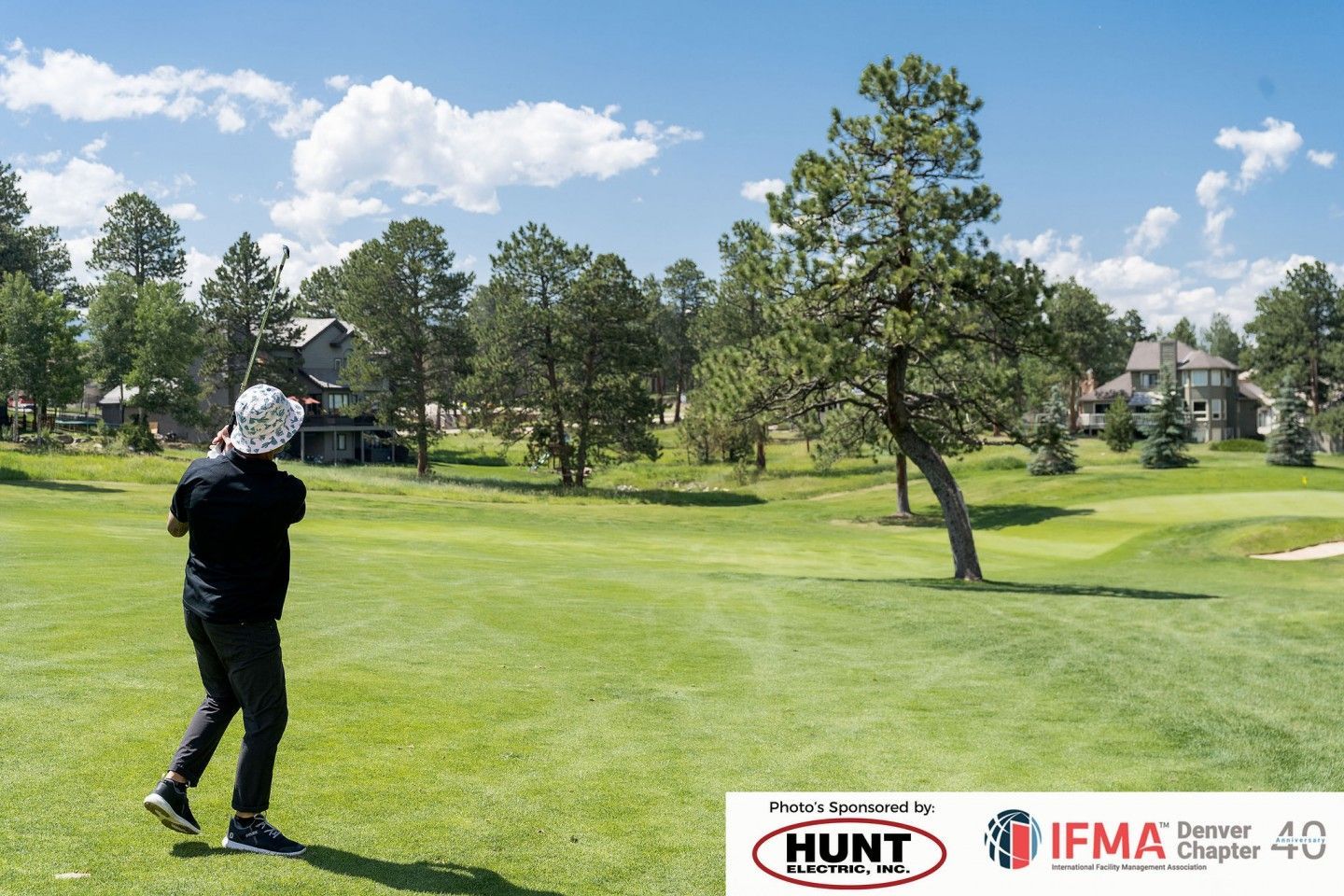 Golfer swinging on a green golf course, with trees and houses in the background. Bright sunny day.