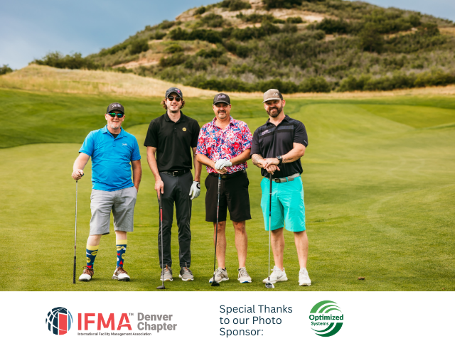 Four men standing on a golf course, posing for a photo. They hold golf clubs in front of a green hill.