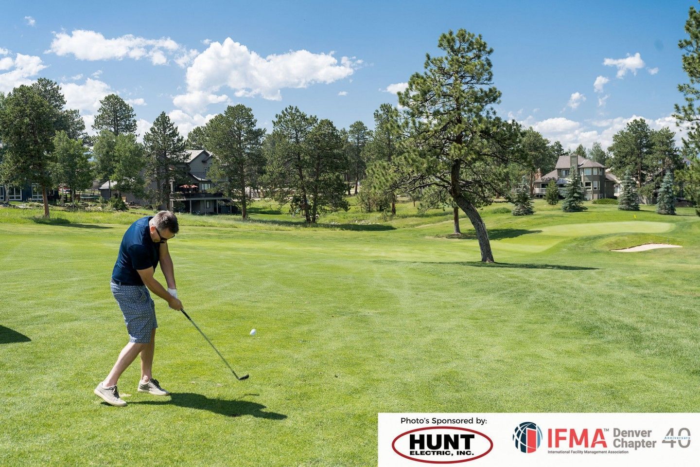 Man swings golf club on a green course under a blue sky, with trees and houses in the background.