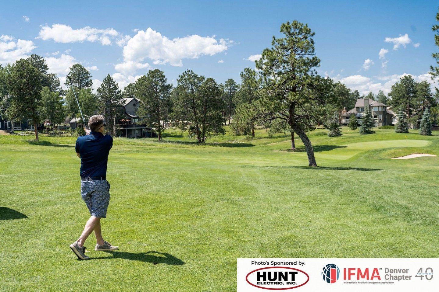 Man swinging a golf club on a green field under a sunny sky, trees and houses in the background.