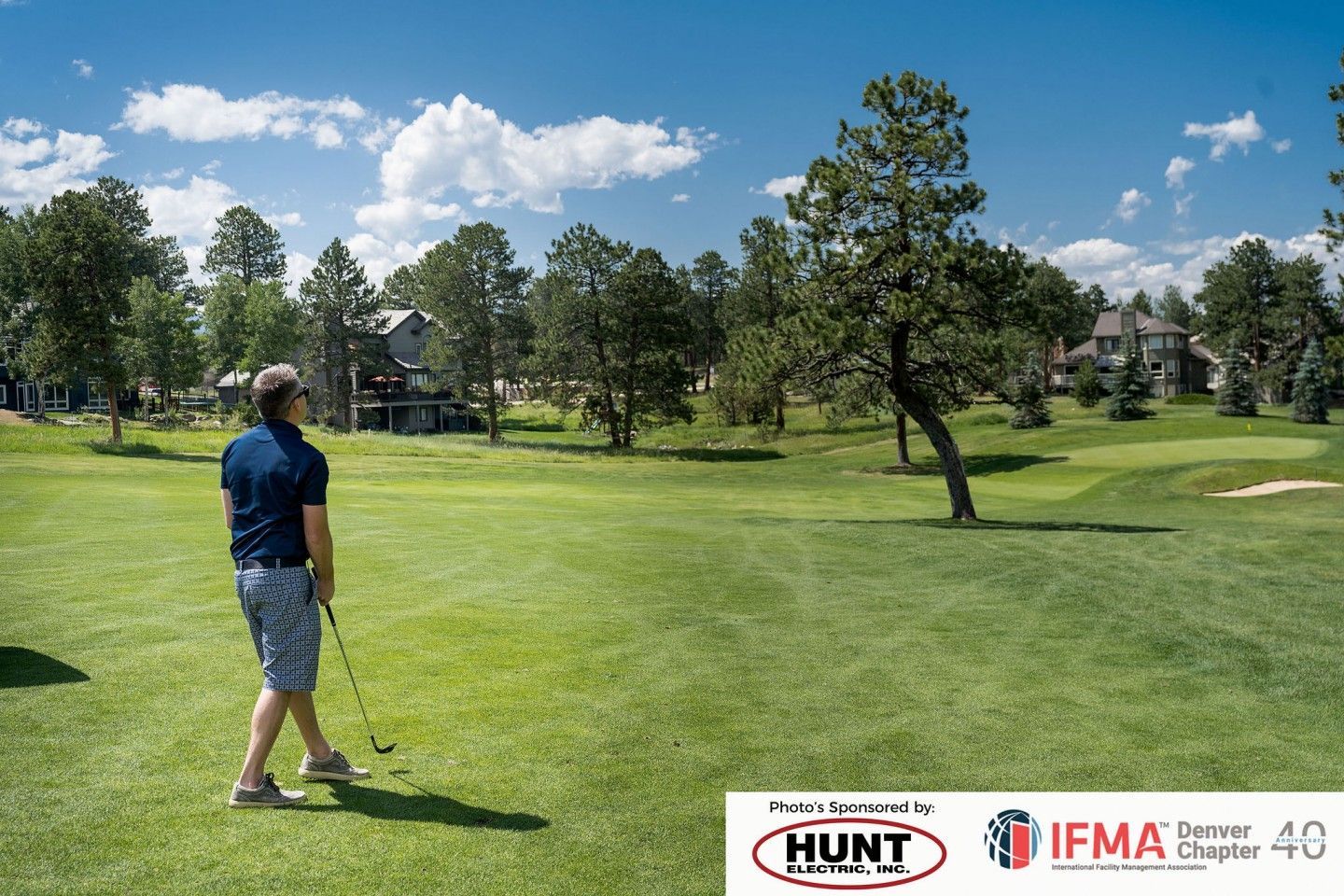 Golfer on a green course, trees and houses in the background on a sunny day.
