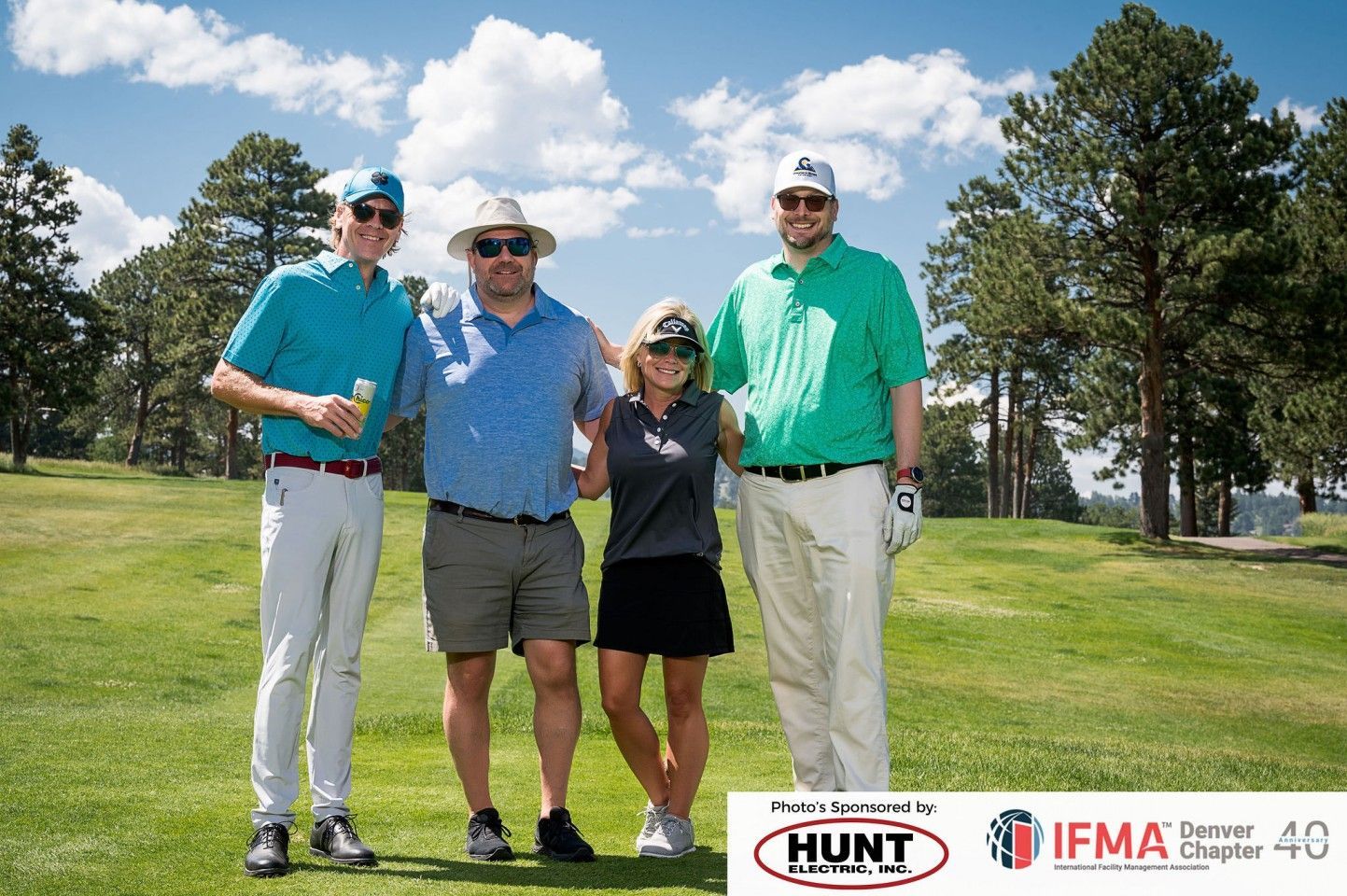 Four people, smiling, posing on a golf course under a blue sky, sponsored by Hunt and IFMA.