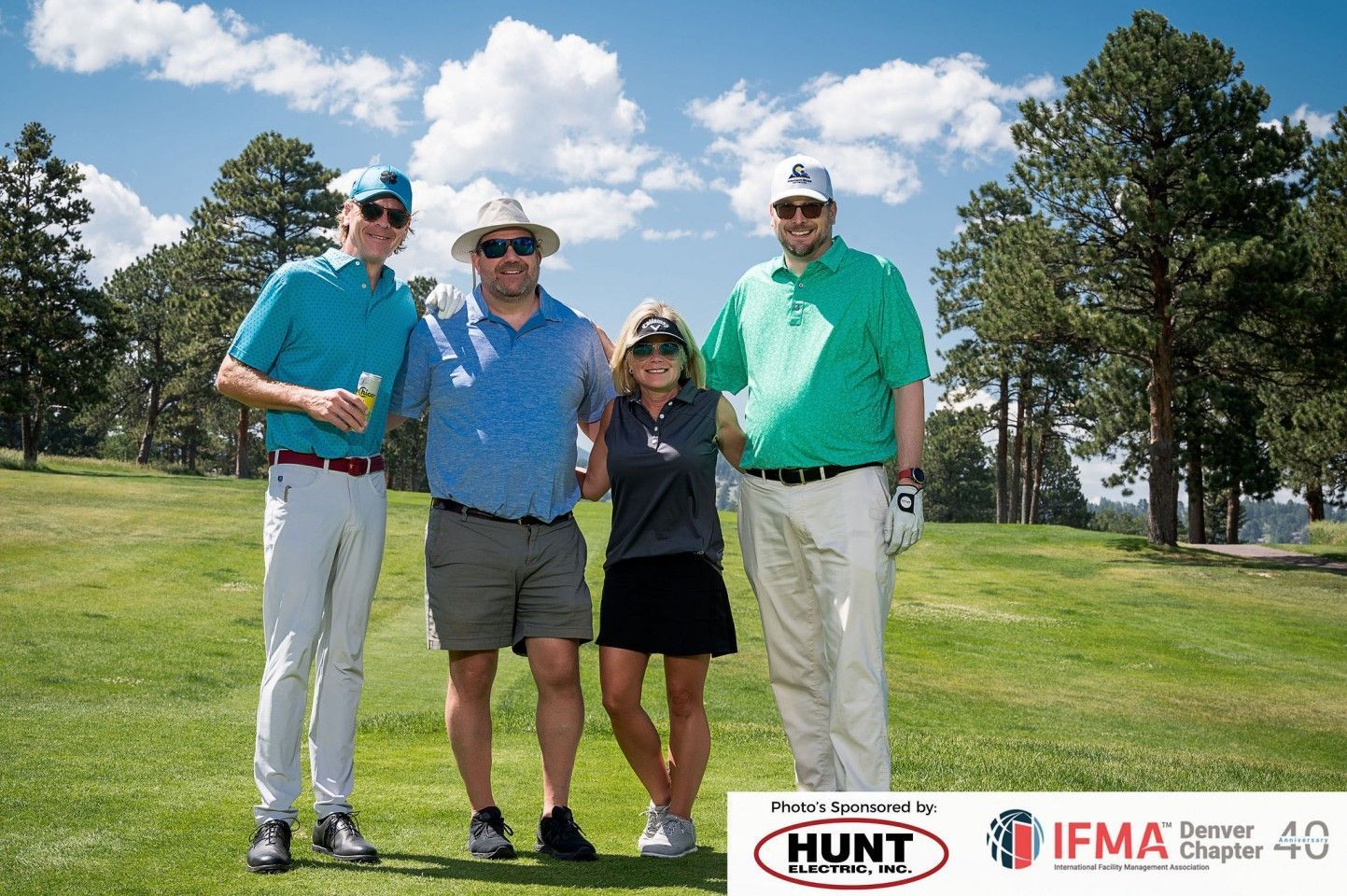 Four people pose on a golf course. Sunny day. Trees in background. Two men wear hats.