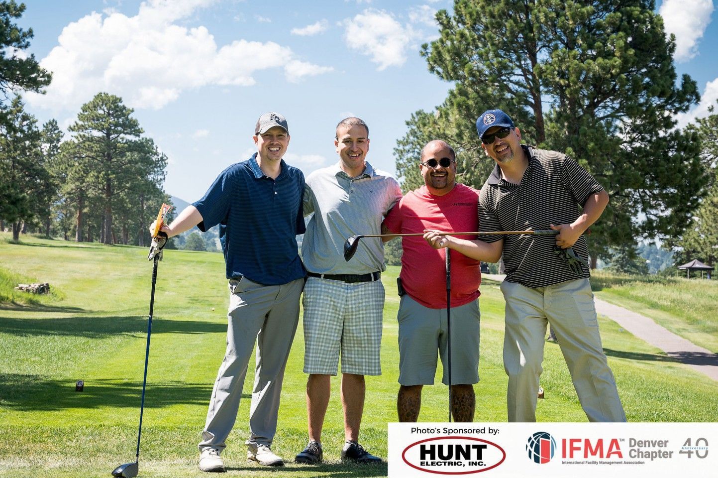 Four men on a golf course smiling, holding clubs. Green grass, trees, and bright sky.