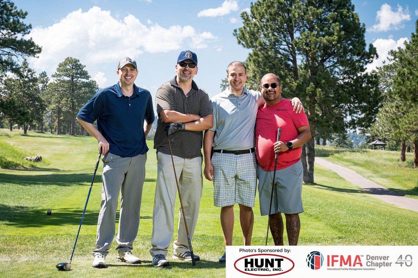 Four men posing on a golf course, holding clubs. Sunny day, green grass, trees.