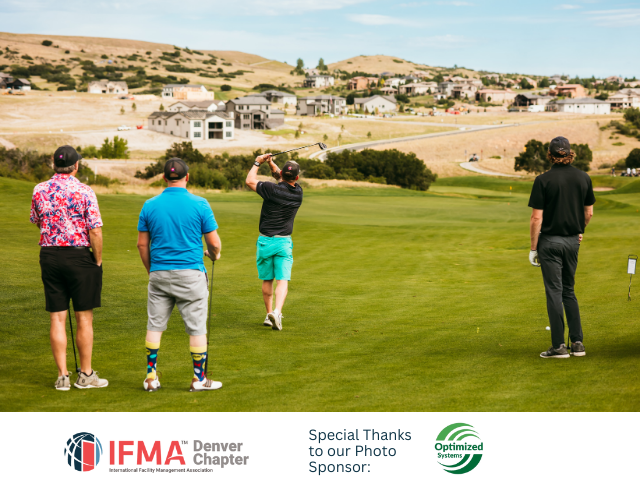 Four golfers on a green; one swings, others watch. Golf course in foreground, houses on a hill in background.