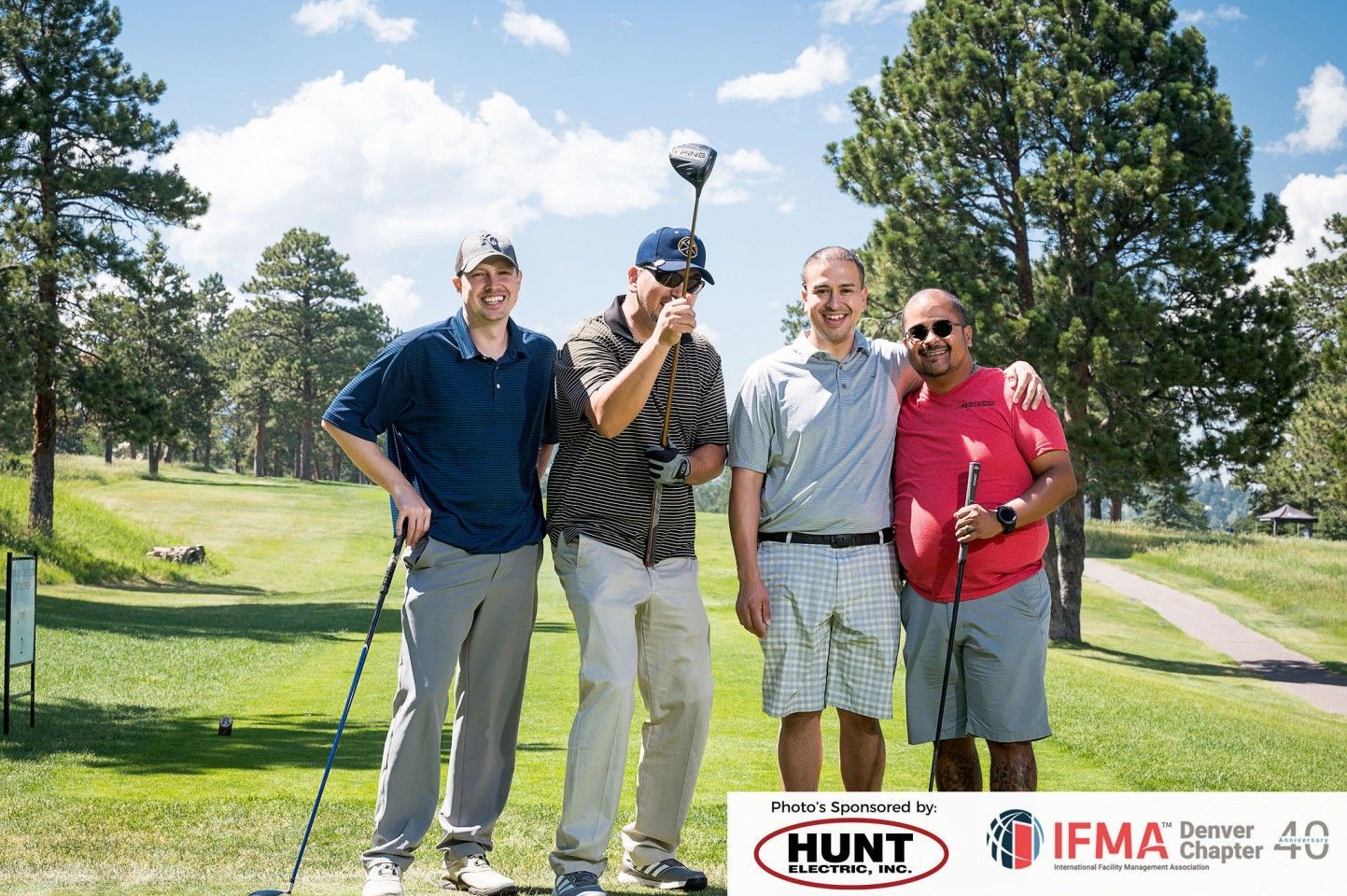 Four men on a golf course smiling, posing with golf clubs, wearing casual clothes. Sunny day.
