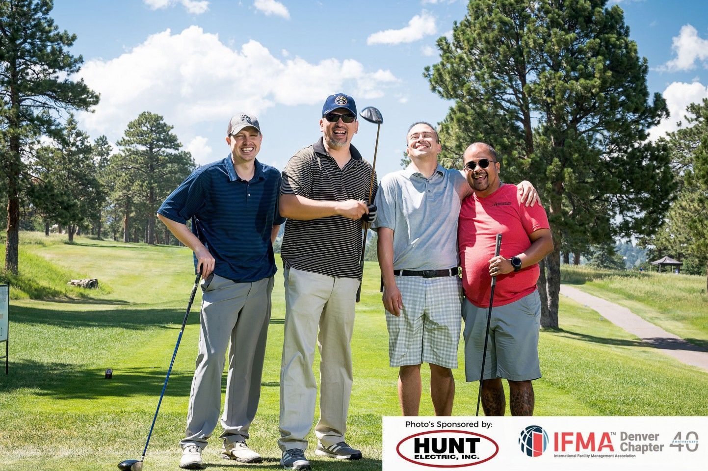 Four men smile on a golf course, posing for a photo. They are in casual golf attire on a sunny day.