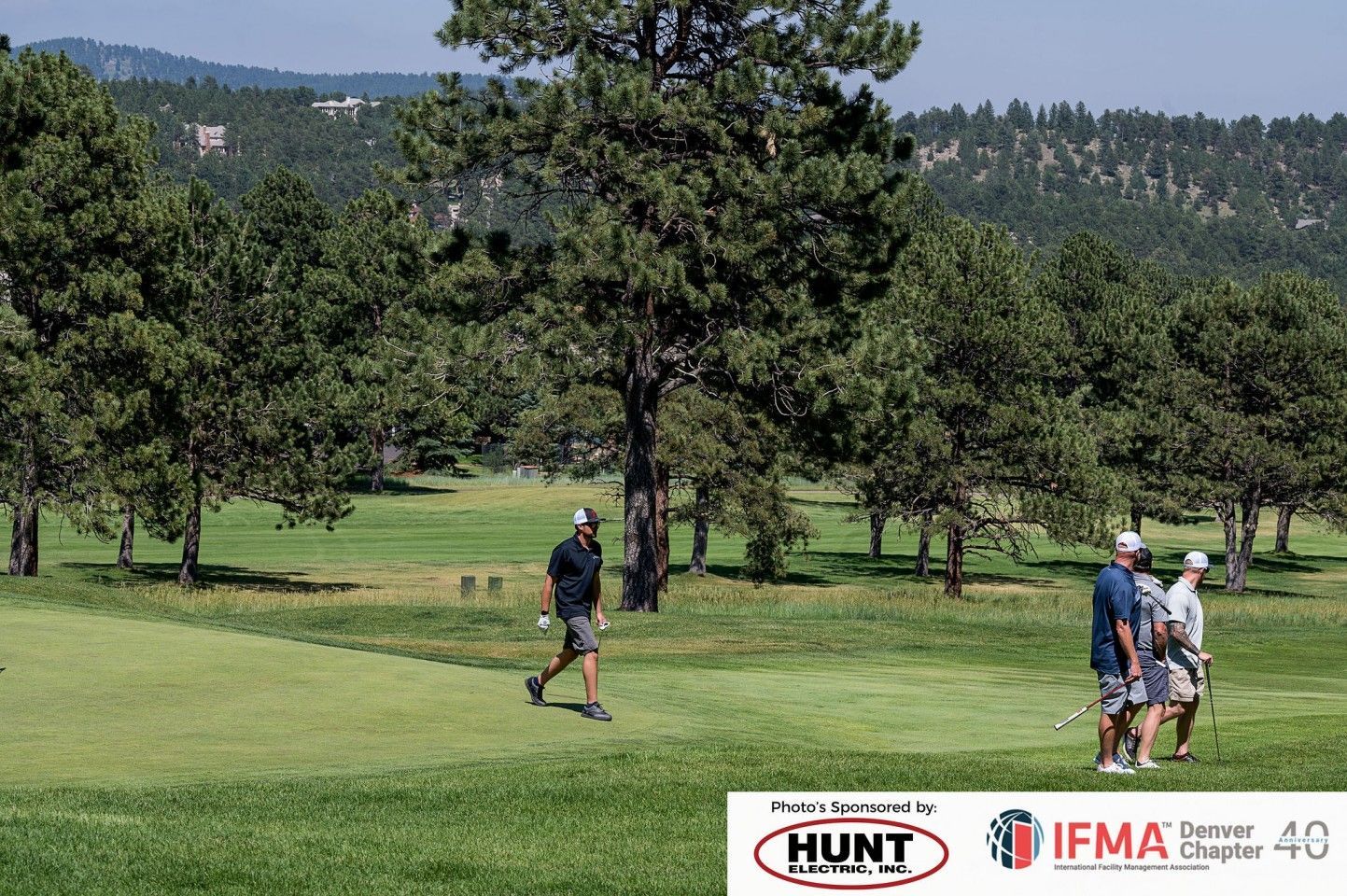 Golfers on a green course with trees, mountains in the background.