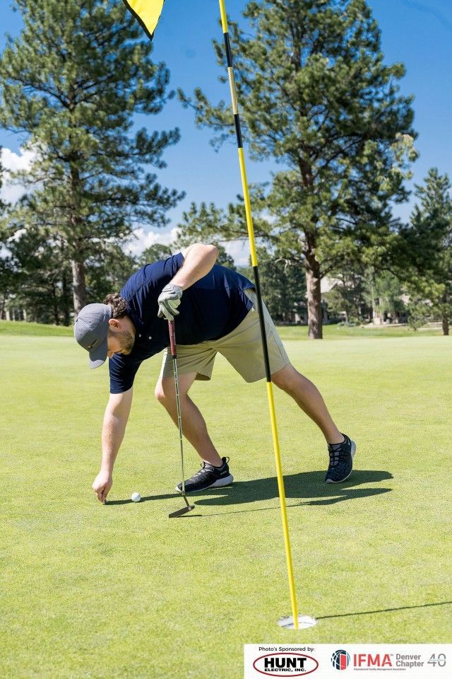 Golfer on green, reaching for ball near flag. Green grass, blue sky, and trees in background.