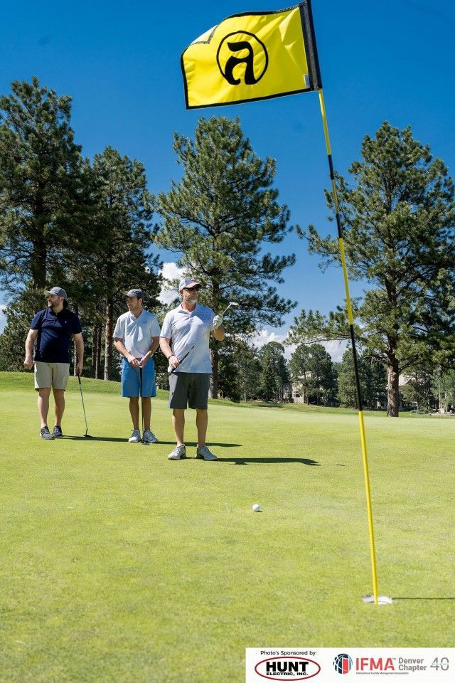 Three golfers on a green, one with club near ball, yellow flag. Blue sky, trees.