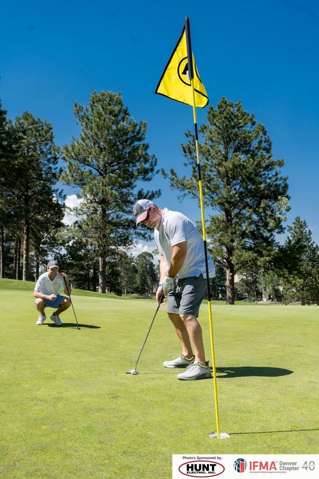 Two golfers on a green, one putting, another watching. Yellow flag marks the hole. Sunny day.