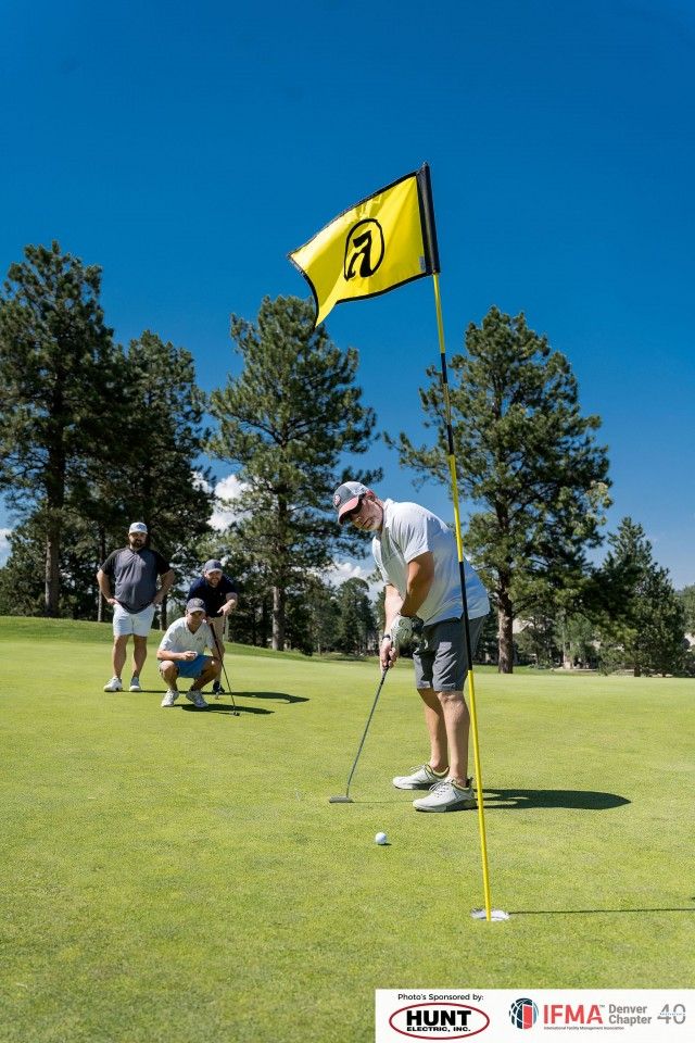 Man putting golf ball on a green, other golfers watch. Yellow flag with logo. Sunny day.