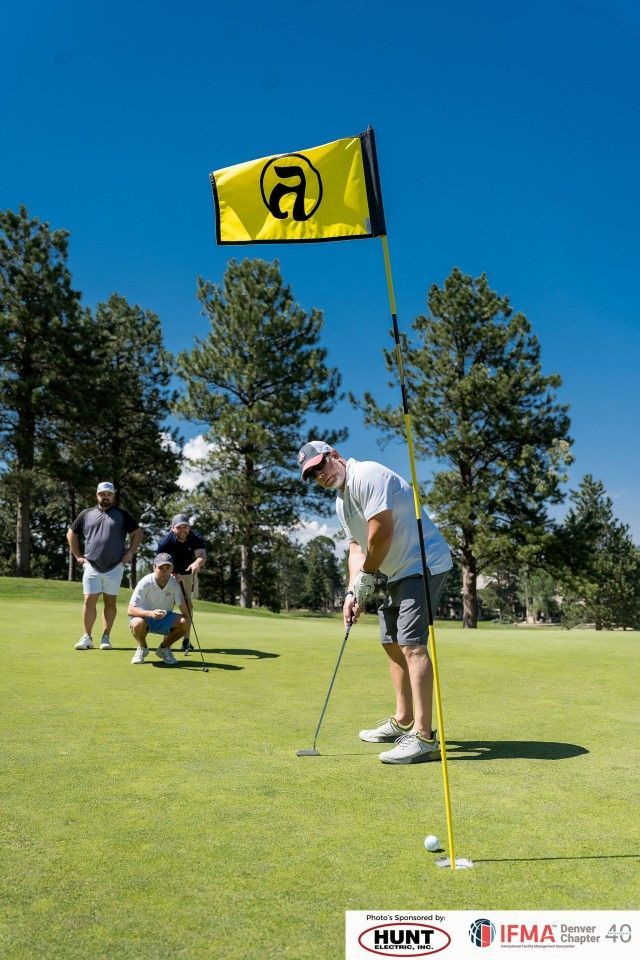Golfer putting on a green, yellow flag in the hole, two observers, sunny day.