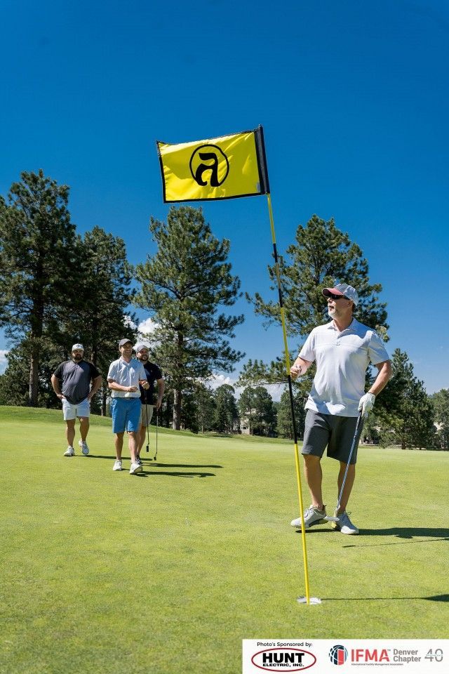 Man on golf course holding flag, others behind, sunny day.