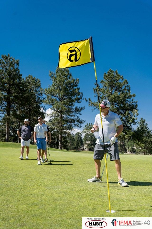 Golfers on green, one holding flagstick near yellow flag, other two behind, trees and blue sky.