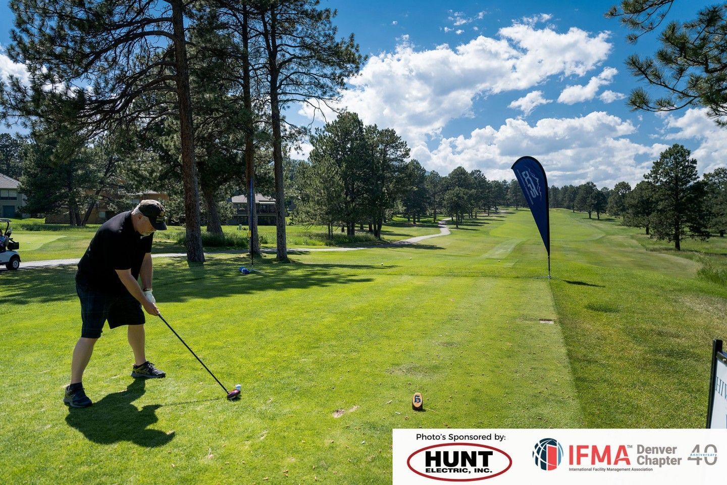 Man in black attire swings golf club on green course; blue banner visible.