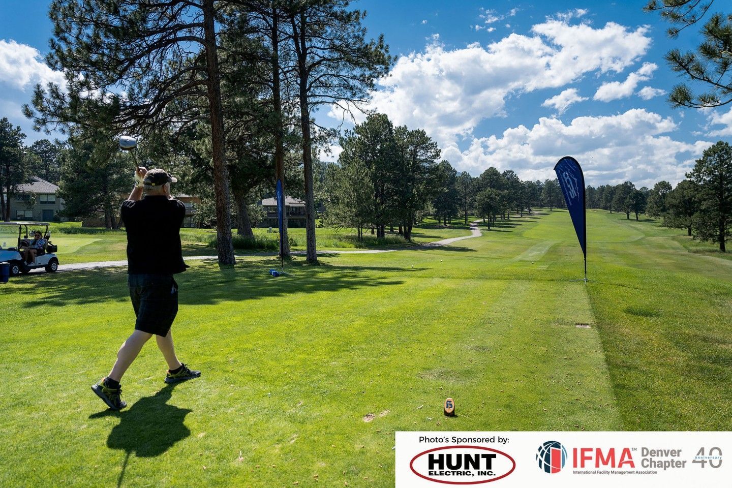 Golfer teeing off on a green golf course under a blue sky, with trees and a golf cart.