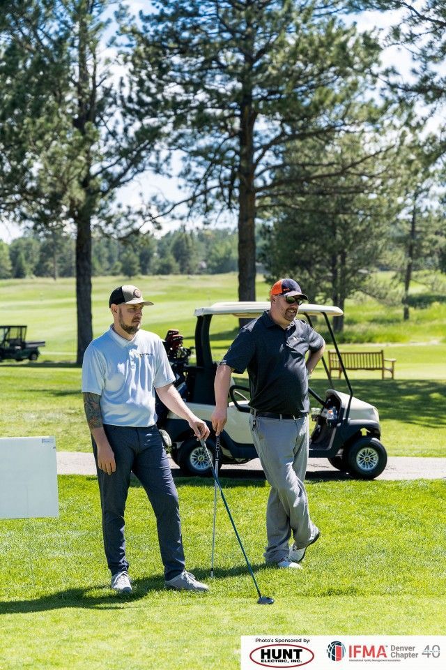Two men on a golf course. One holds a club, the other wears a cap, standing near a golf cart.