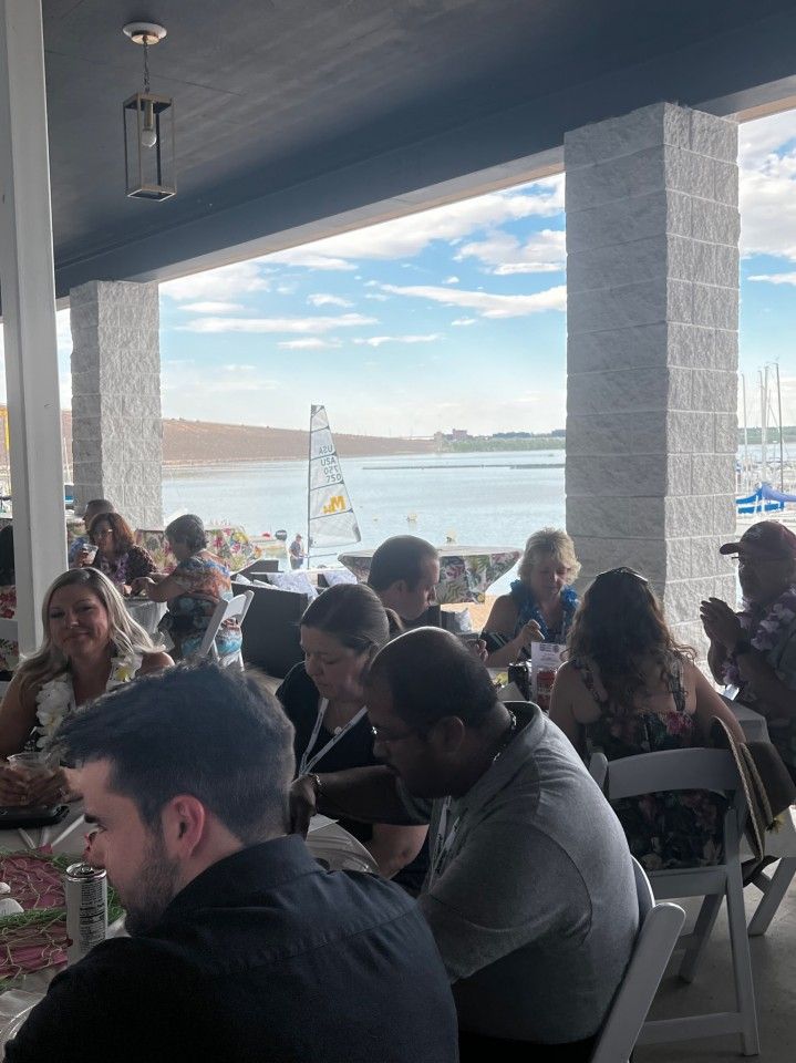 People seated at tables on a patio overlooking a lake, possibly at an event.