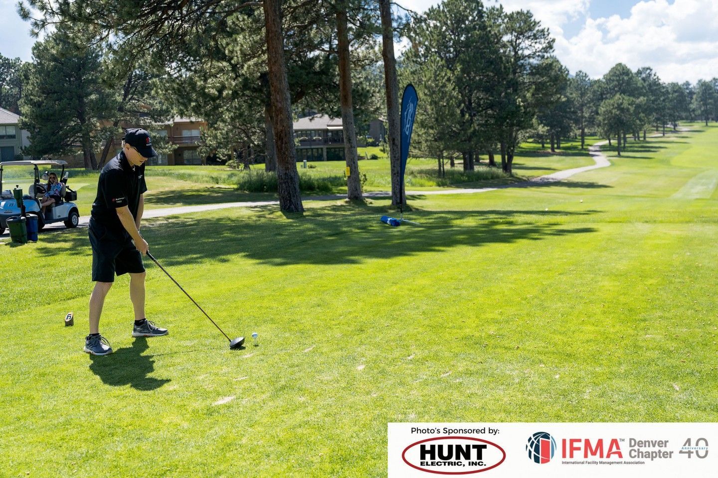 Golfer teeing off on a green golf course, sunny day. Trees and golf carts in background.