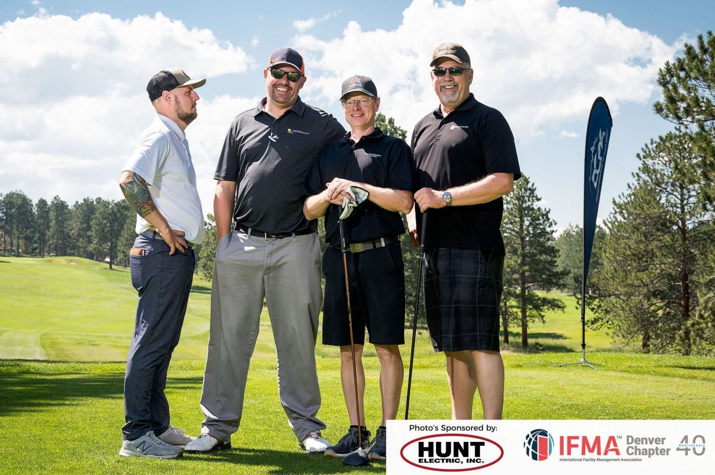 Four men on a golf course pose for a photo. They wear golf attire. Green grass and trees are in the background.