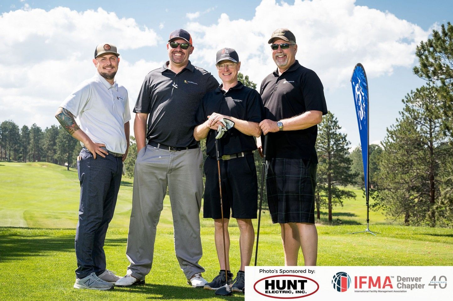 Four men on a golf course, posing for a photo. They are smiling, holding golf clubs. Sunny day.