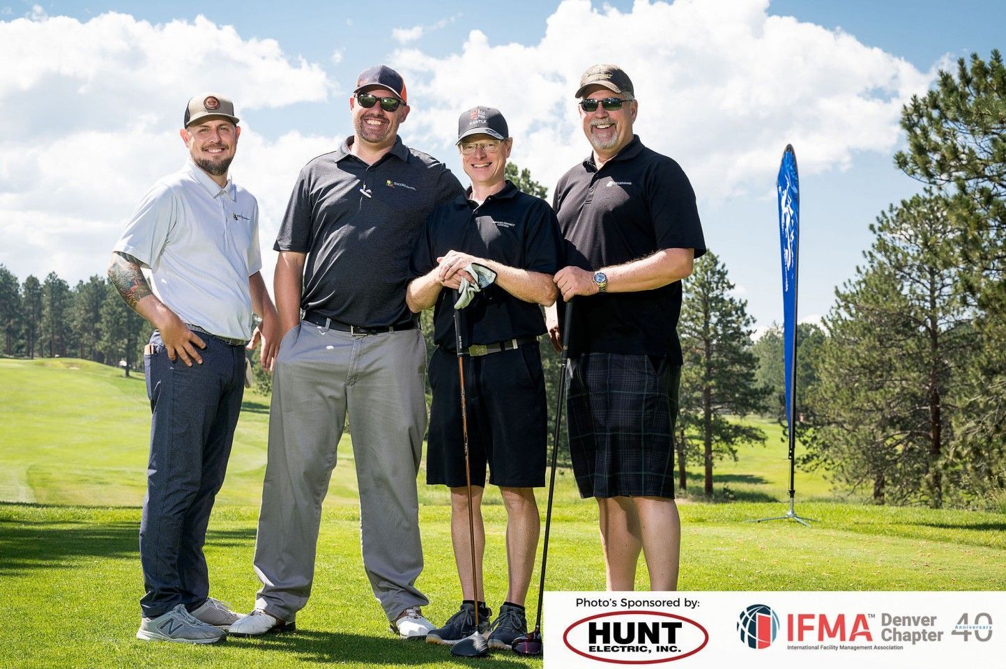 Four men posing on a golf course, smiling with golf clubs. Sunny day, green grass, blue sky.