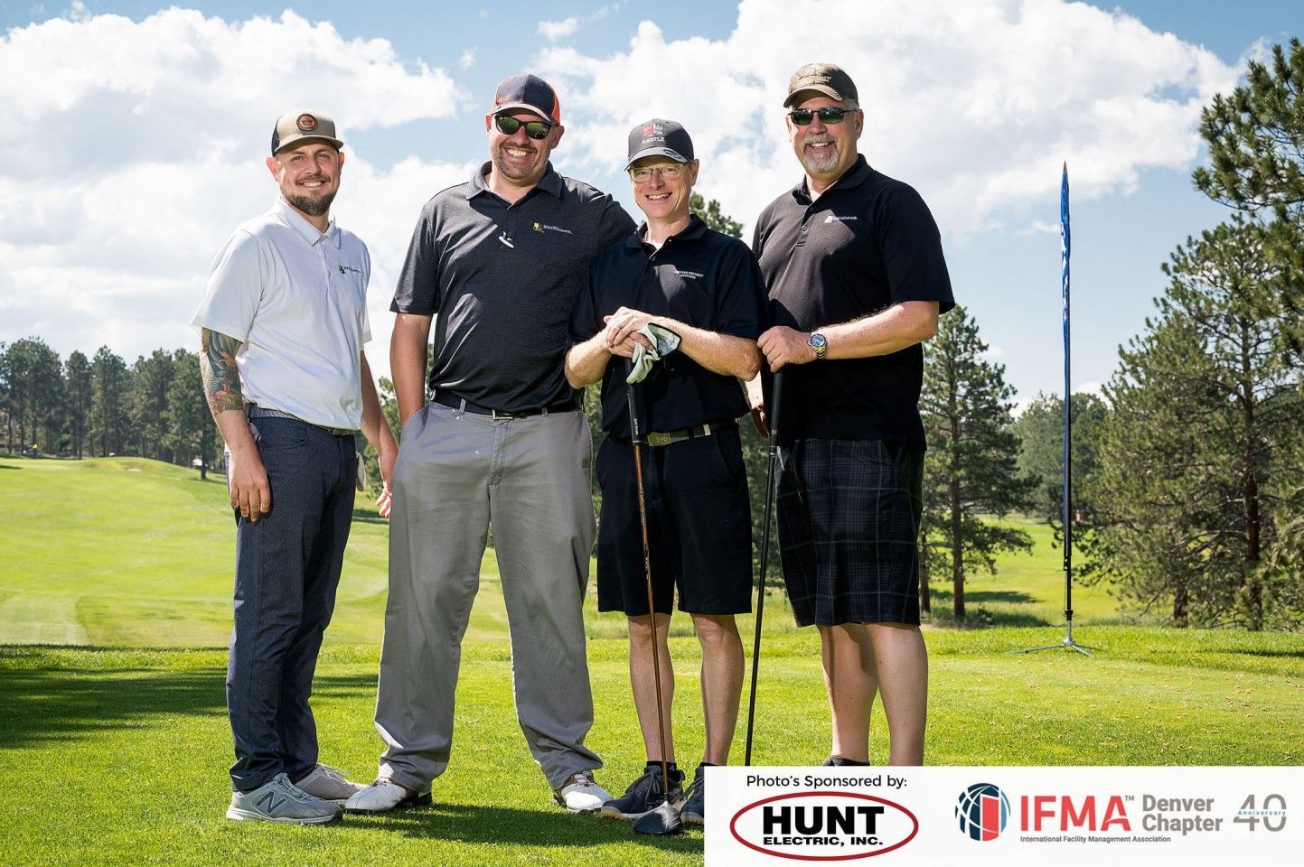 Four men on a golf course pose for a photo. They are wearing hats and golf attire.