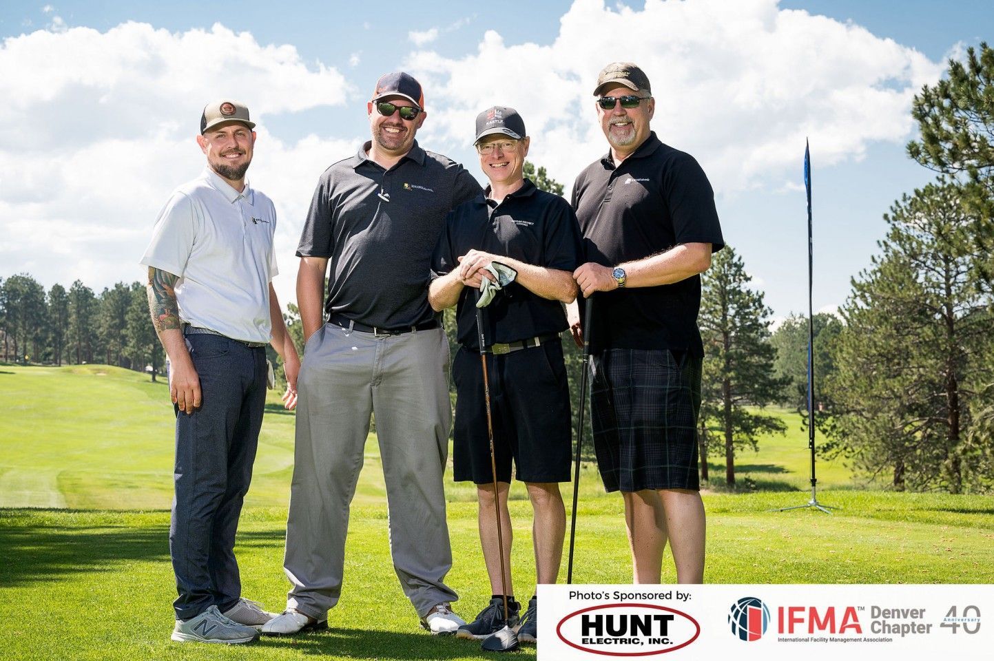 Four men on a golf course pose with clubs. Sunny day, green grass, blue sky. Logos at bottom.
