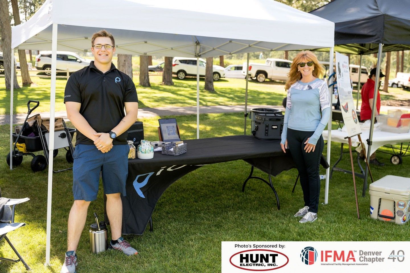Two people stand at a booth under a canopy in a park.