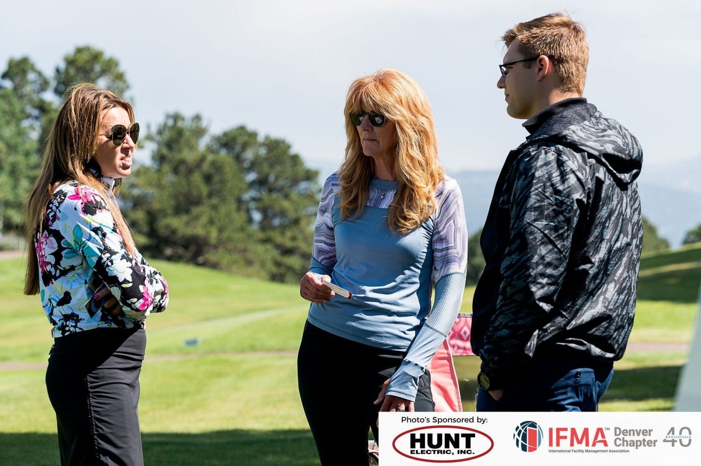 Three people on a golf course talking: woman in blue shirt, another in patterned shirt, and a man in a jacket.