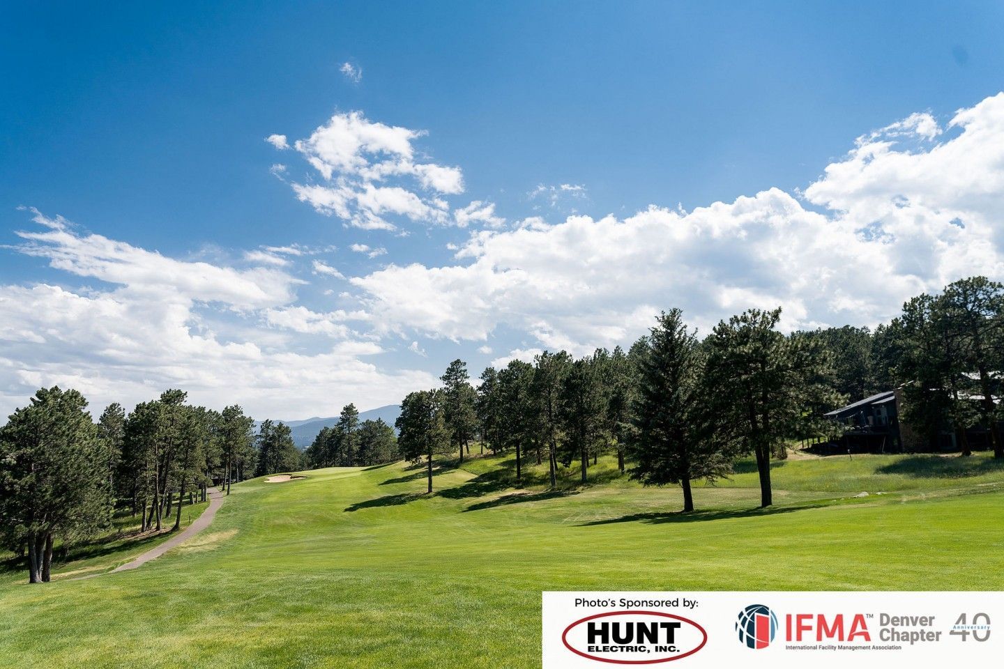 Green golf course under a blue sky with fluffy clouds and trees.