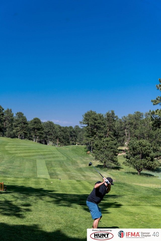 Golfer swings club on a sunny golf course with green grass, trees, and a blue sky.