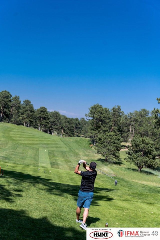 Golfer swinging a club on a sunny green golf course. Bright blue sky, trees in the background.