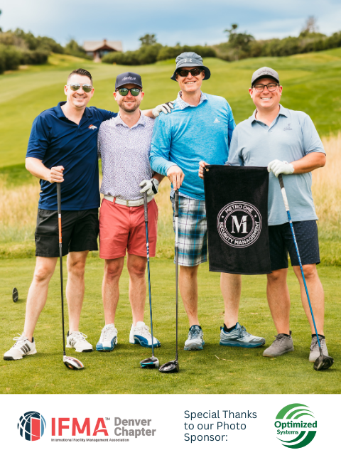 Four men on a golf course pose with clubs, holding a flag. Green grass and blue sky.