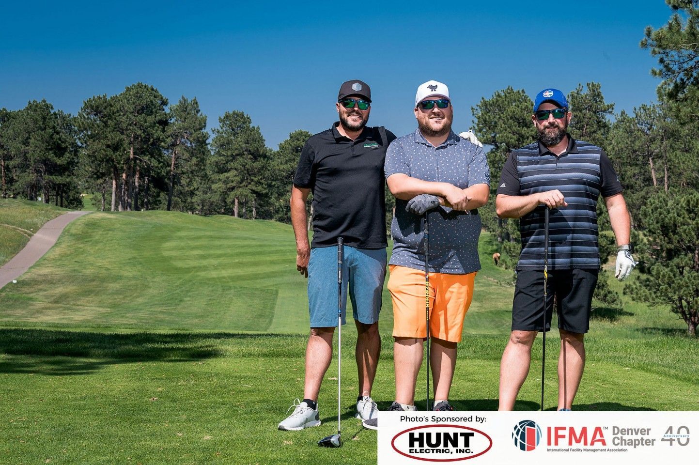 Three men posing on a golf course, holding clubs. Green grass, blue sky, and trees in the background.