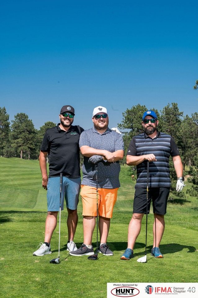 Three men on a golf course, posing with clubs. Blue sky, green grass.