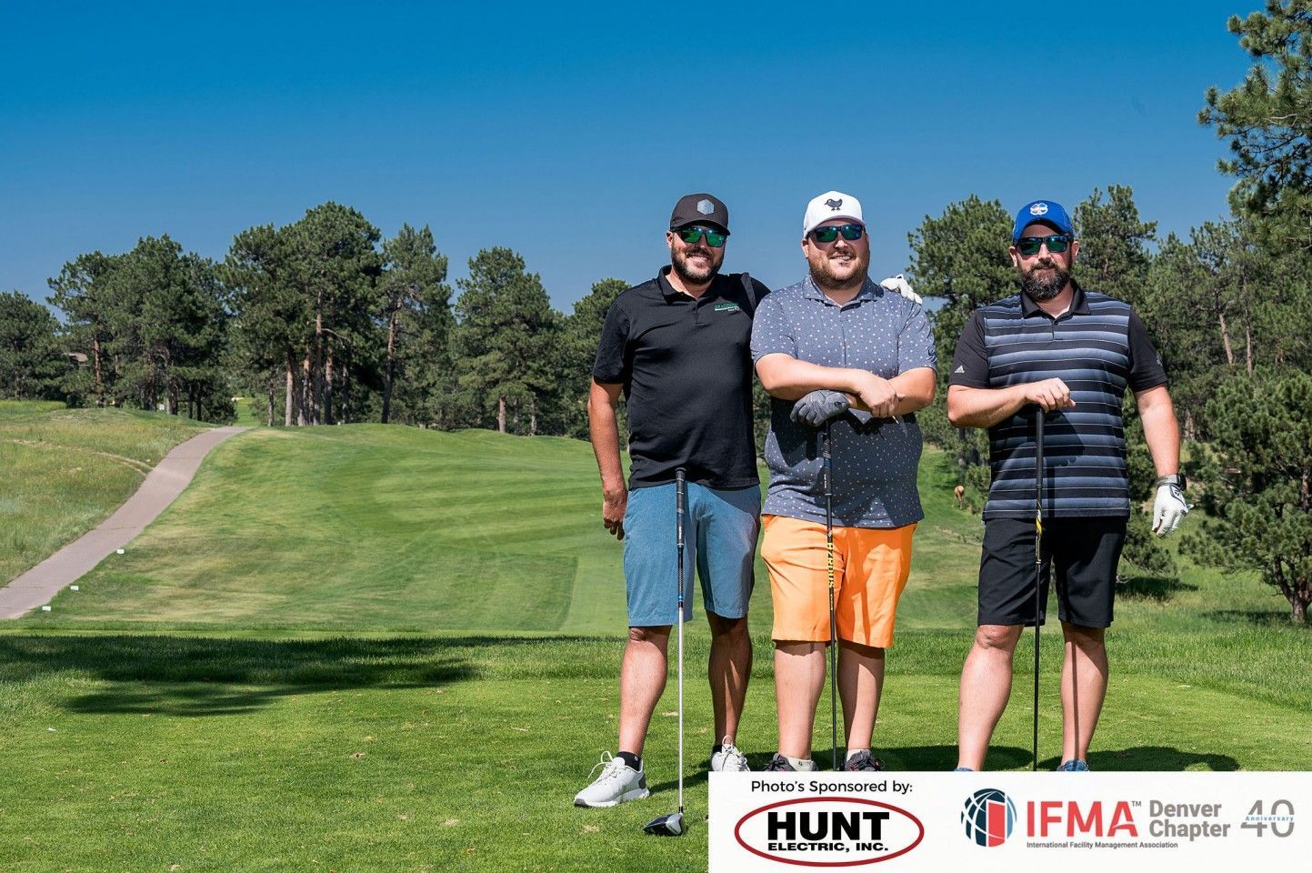 Three men with golf clubs on a green course, posing with a blue sky background.
