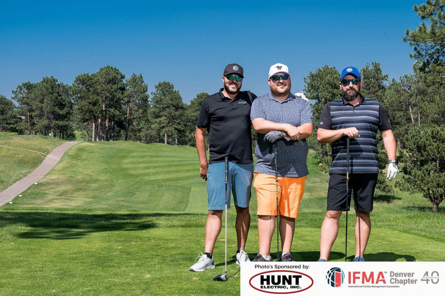 Three men on a golf course, smiling, holding clubs, wearing hats and casual clothes. Sunny day.