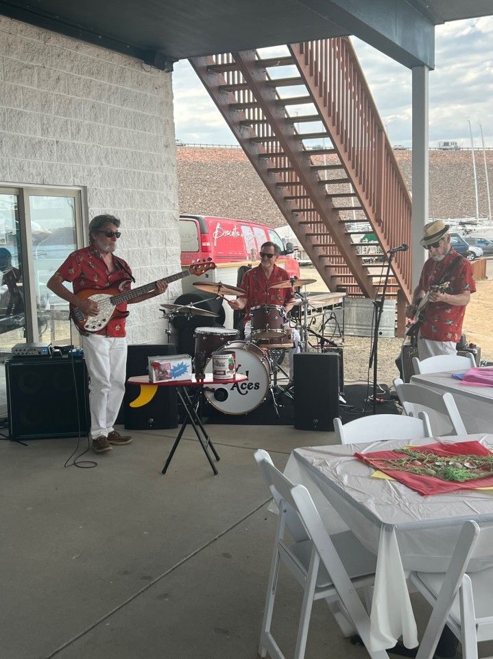 A band performs outdoors. Three musicians play instruments wearing red shirts and white pants.