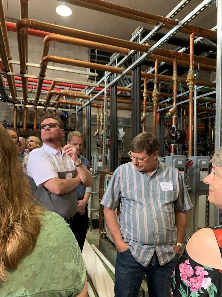 Group of people looking at copper pipes in a mechanical room; man in blue shirt is smiling.