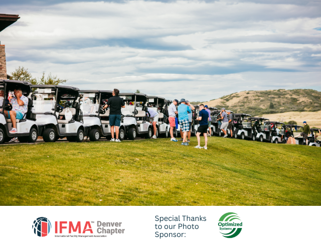 Golfers stand near golf carts on a green hill with a building in the background. Cloudy sky.