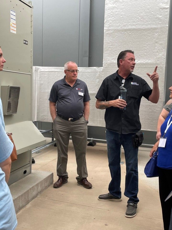 Two men and a group in a mechanical room; one man gestures, the other looks on.