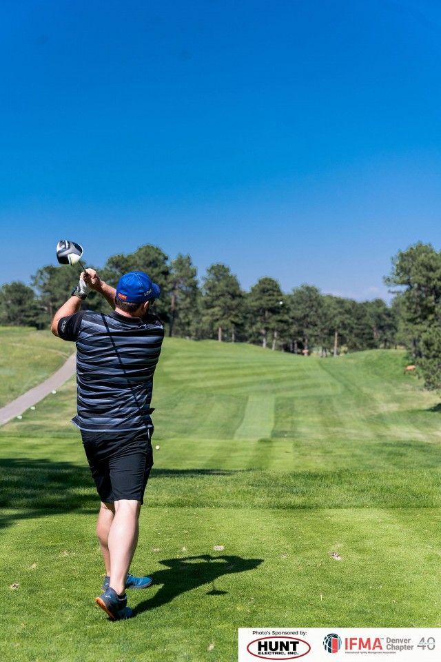 Golfer in blue hat swings club on a green golf course under a clear blue sky.