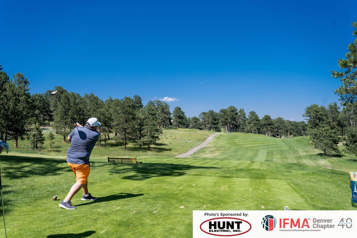 Golfer swings on a green golf course under a blue sky.