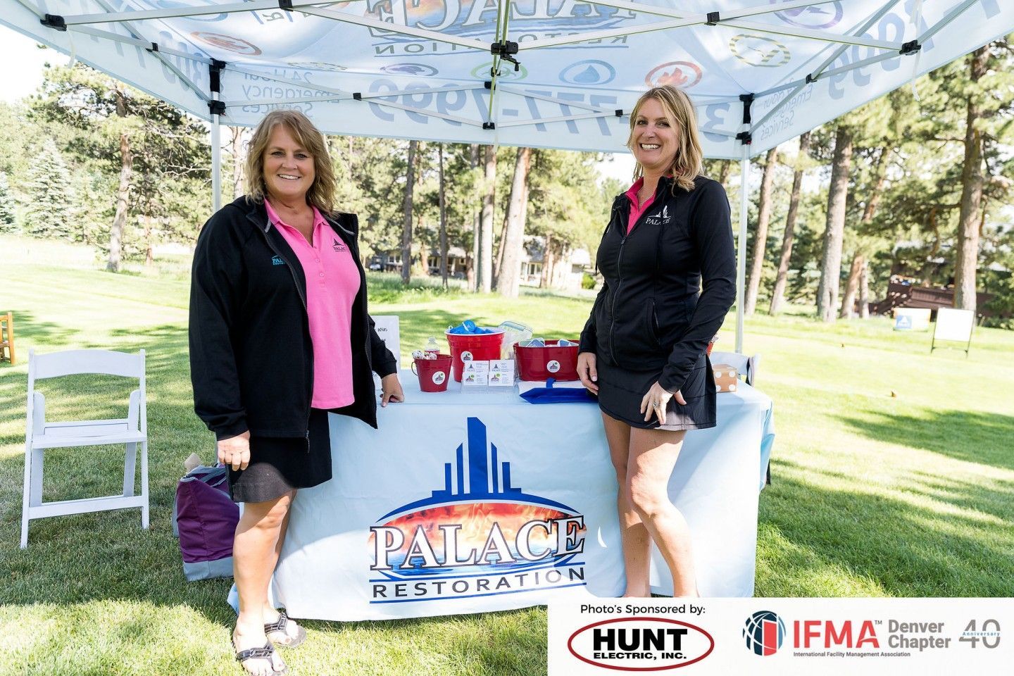 Two women stand at a booth promoting Palace Restoration; outdoor setting with gifts and trees.