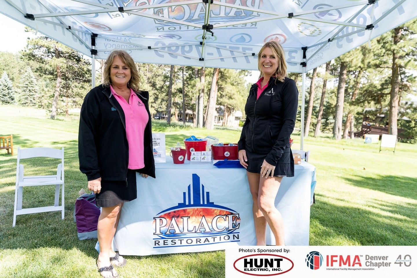 Two women at a Palace Restoration booth. One in pink shirt, other in black. Table with logo, outdoors.