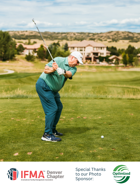 Golfer swinging club on a green course, wearing a white hat, green shirt, and blue pants. Buildings and hills in background.